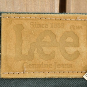 May include: A brown leather patch with the text "Since 1889" and "Lee Genuine Jeans" in a stylized font. The patch is sewn onto a dark green fabric.