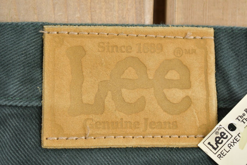 May include: A brown leather patch with the text "Since 1889" and "Lee Genuine Jeans" in a stylized font. The patch is sewn onto a dark green fabric.