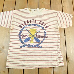 May include: Cream-colored, striped t-shirt with a nautical design. The shirt features red horizontal stripes and a circular emblem with the words "Regatta Gold," "Skipper," and "Helmsman." The emblem includes crossed oars and a trophy.