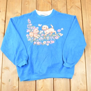 May include: A bright blue sweatshirt with a white collar. The front displays a floral design with two puppies and a butterfly. The puppies are light brown and cream-coloured, surrounded by pink and purple flowers.