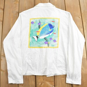 May include: White long-sleeved shirt with a colourful fish design on the back. The fish is blue, yellow, and white with a green and blue background. The design is framed in yellow.