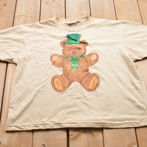 May include: A beige t-shirt with a brown teddy bear wearing a green hat and a green bow tie.