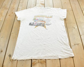 May include: A white short-sleeve t-shirt featuring a graphic print of a race car on a yellow star background. The text on the graphic reads Knoxville and includes the word Knoxville, Iowa, with a small checkered flag design above the car.
