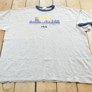 Puede incluir: Camiseta gris con cuello y ribetes de manga azules. La camiseta presenta un gr&aacute;fico de horizonte con el texto "US Open New York 99" y el logotipo de Fila. La camiseta est&aacute; sobre una superficie de madera.