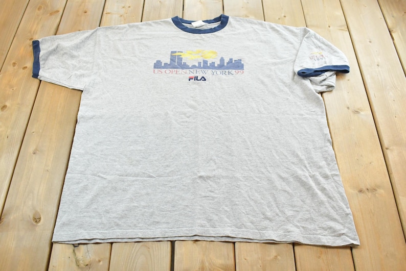 Puede incluir: Camiseta gris con cuello y ribetes de manga azules. La camiseta presenta un gr&aacute;fico de horizonte con el texto "US Open New York 99" y el logotipo de Fila. La camiseta est&aacute; sobre una superficie de madera.