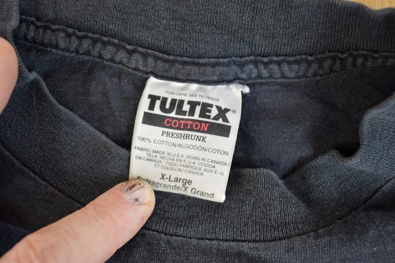 May include: Black T-shirt with a white Tultex tag that reads "100% Cotton/Algodon/Coton Preshrunk" and "Fabric Made in USA Sewn in Canada/En Canada Tissu Fabrique aux E.U./Tela Mecho en EUA Cosida et Cousu au Canada" and "X-Large/Extra Grande/X Grand".