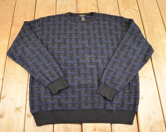 May include: A crew neck jumper with a woven pattern in shades of blue, black, and grey. The cuffs, neckline, and hem are solid black. The jumper is made of a soft, textured material.