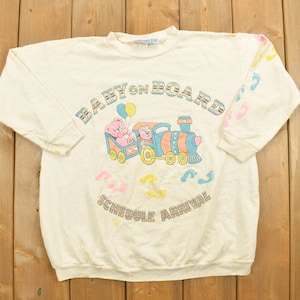 May include: A white long-sleeved sweatshirt with a graphic of a train with two teddy bears on board. The text "Baby on Board" is above the train and "Schedule Arrival" is below the train. The sleeves have a pattern of pink and blue footprints.