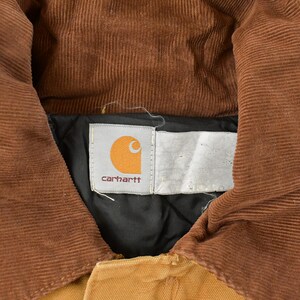 Puede incluir: Primer plano del cuello de una chaqueta Carhartt. El cuello es de pana marr&oacute;n y la chaqueta es de color canela. El logotipo de Carhartt es visible en una etiqueta blanca. La chaqueta est&aacute; hecha de un material de trabajo duradero.