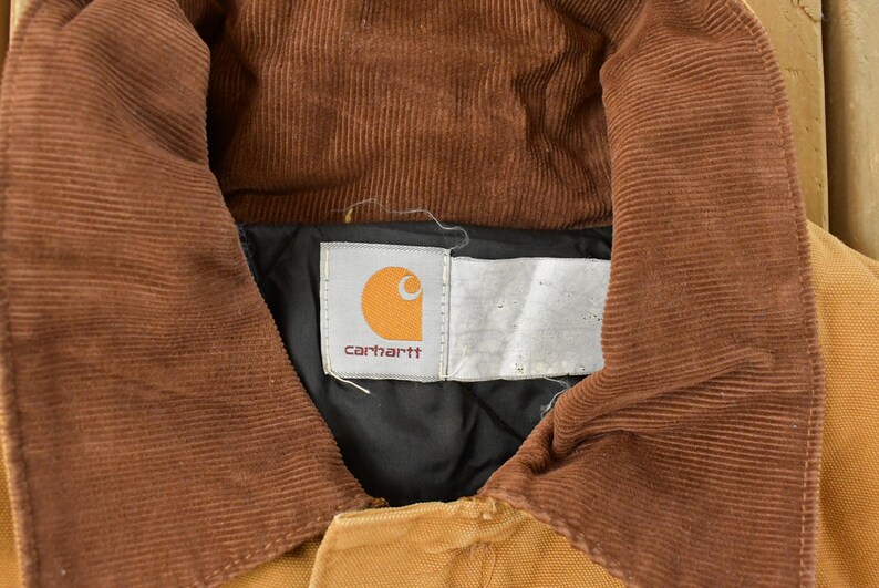 Puede incluir: Primer plano del cuello de una chaqueta Carhartt. El cuello es de pana marr&oacute;n y la chaqueta es de color canela. El logotipo de Carhartt es visible en una etiqueta blanca. La chaqueta est&aacute; hecha de un material de trabajo duradero.