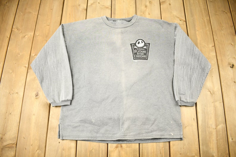 Puede incluir: Sudadera gris de manga larga con mangas acanaladas. La parte delantera presenta un gr&aacute;fico de "Pesadilla antes de Navidad" con Jack Skellington. La camisa tiene cuello redondo y un ajuste relajado.