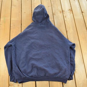 K&ouml;nnte beinhalten: Ein marineblauer Kapuzenpullover, von hinten betrachtet, liegt auf einer Holzoberfl&auml;che. Der Pullover hat eine Kapuze mit Kordelzug, lange &Auml;rmel und gerippte B&uuml;ndchen und Saum. Der Stoff wirkt weich und leicht abgenutzt.