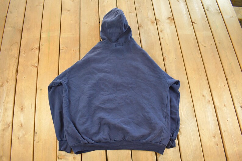 K&ouml;nnte beinhalten: Ein marineblauer Kapuzenpullover, von hinten betrachtet, liegt auf einer Holzoberfl&auml;che. Der Pullover hat eine Kapuze mit Kordelzug, lange &Auml;rmel und gerippte B&uuml;ndchen und Saum. Der Stoff wirkt weich und leicht abgenutzt.