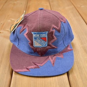 May include: A blue and burgundy baseball cap with a New York Rangers logo embroidered on the front. The cap has a jagged, zig-zag pattern around the logo.
