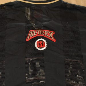 Op de afbeelding: Zwart mesh shirt met verticale strepen en een gouden kraag. Het shirt heeft het "AVIREX" logo in rood en wit, met een "USA" embleem eronder. Het shirt is waarschijnlijk vintage.
