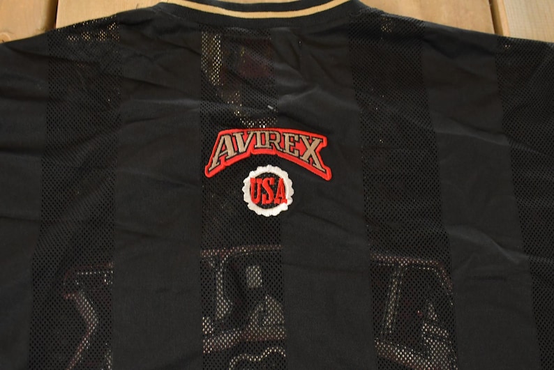 Op de afbeelding: Zwart mesh shirt met verticale strepen en een gouden kraag. Het shirt heeft het "AVIREX" logo in rood en wit, met een "USA" embleem eronder. Het shirt is waarschijnlijk vintage.