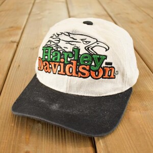 Op de afbeelding: Een vintage Harley-Davidson baseball cap. De pet heeft een witte kroon en een zwarte klep. Het Harley-Davidson-logo is geborduurd in groen en oranje, met een adelaarsafbeelding boven de tekst.