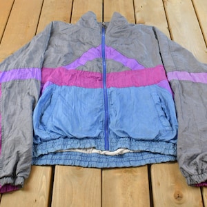 Puede incluir: Una chaqueta cortavientos de manga larga con un diseño de bloques de color en tonos gris, azul brillante y magenta. La chaqueta cuenta con una cremallera frontal azul de longitud completa, cuello alto, puños elásticos y dobladillo elástico.