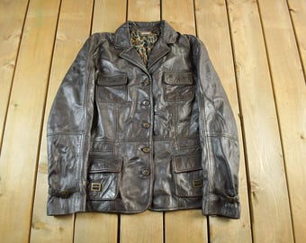 May include: A brown leather jacket with four front pockets, a collared neckline, and button closures. The jacket has a leopard print lining and buckle details on the sleeves. The jacket is displayed on a wooden surface.