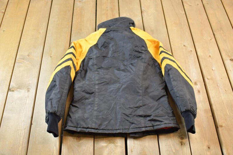 May include: Black jacket with yellow stripes on the sleeves. The jacket has a zipper closure and a high collar.