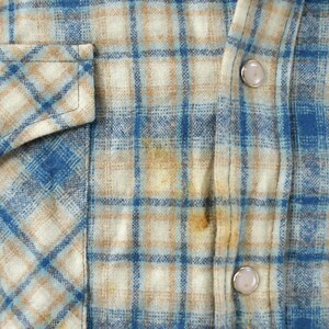 May include: Close-up of a blue, tan, and white plaid shirt. The shirt has a button-down front, a chest pocket with a silver snap, and silver snaps on the cuffs. The fabric appears to be a soft flannel.