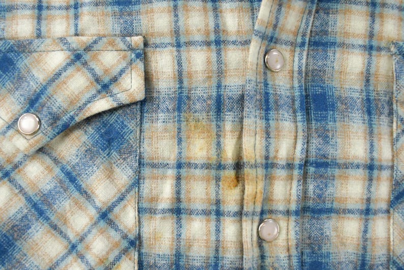 May include: Close-up of a blue, tan, and white plaid shirt. The shirt has a button-down front, a chest pocket with a silver snap, and silver snaps on the cuffs. The fabric appears to be a soft flannel.