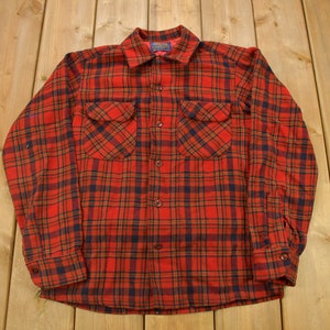 May include: A red, black and orange plaid flannel shirt with long sleeves and a button-up front.