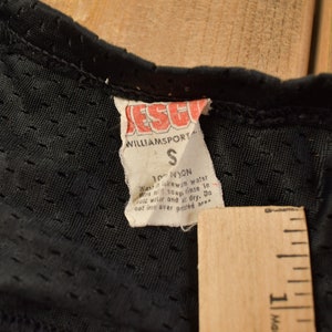 Op de afbeelding: Close-up van een zwart label met witte tekst die luidt: "ESCORT WILLIAMSPORT S 100% NYLON Was in lauw water met milde zeep. Spoel in koud water en laat aan de lucht drogen. Niet strijken of chemisch reinigen. Niet vouwen over het bedrukte gebied."