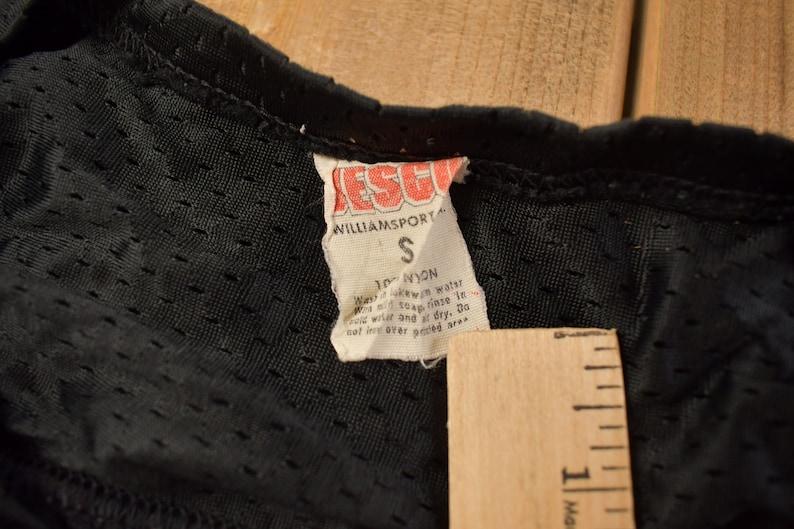 Op de afbeelding: Close-up van een zwart label met witte tekst die luidt: "ESCORT WILLIAMSPORT S 100% NYLON Was in lauw water met milde zeep. Spoel in koud water en laat aan de lucht drogen. Niet strijken of chemisch reinigen. Niet vouwen over het bedrukte gebied."
