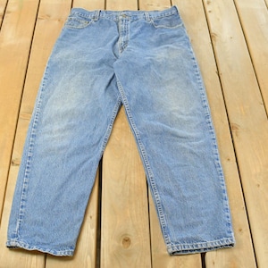 Puede incluir: Vaqueros de mezclilla azul claro. Los vaqueros est&aacute;n colocados sobre una superficie de madera. Los vaqueros tienen un dise&ntilde;o cl&aacute;sico de cinco bolsillos, cierre de bot&oacute;n y cremallera, y trabillas. La mezclilla tiene un aspecto descolorido.