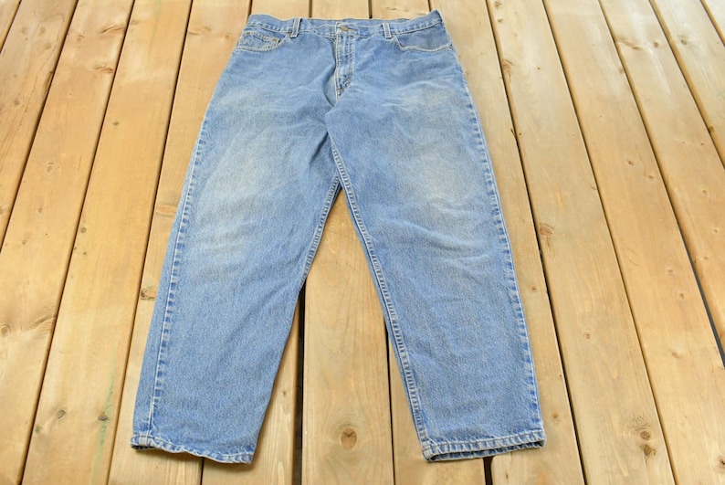 Puede incluir: Vaqueros de mezclilla azul claro. Los vaqueros est&aacute;n colocados sobre una superficie de madera. Los vaqueros tienen un dise&ntilde;o cl&aacute;sico de cinco bolsillos, cierre de bot&oacute;n y cremallera, y trabillas. La mezclilla tiene un aspecto descolorido.