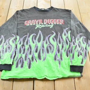 May include: A long-sleeved, gray shirt with a green flame design at the bottom and sleeves. The shirt features the text "GRAVE DIGGER MONSTER TRUCK RACING" in red and green across the chest. The shirt has black cuffs and a black collar.