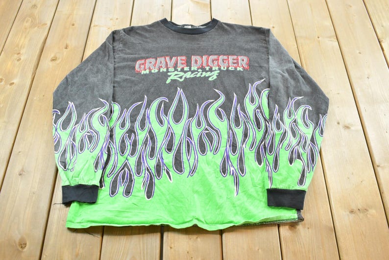 May include: A long-sleeved, gray shirt with a green flame design at the bottom and sleeves. The shirt features the text "GRAVE DIGGER MONSTER TRUCK RACING" in red and green across the chest. The shirt has black cuffs and a black collar.