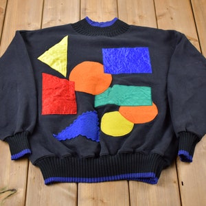Puede incluir: Sudadera negra con un diseño geométrico colorido. El diseño presenta una variedad de formas, incluyendo círculos, cuadrados y triángulos, en tonos de rojo, naranja, amarillo, verde y azul.
