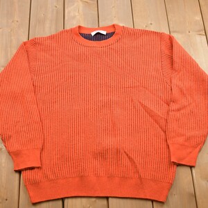 May include: A bright orange knitted jumper with a ribbed texture. The jumper has a crew neck and long sleeves.