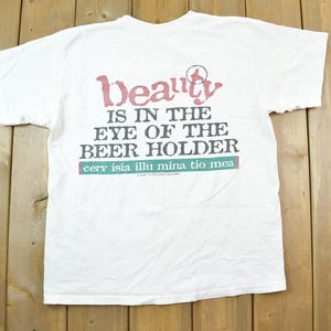 Puede incluir: Camiseta blanca con la frase "Beauty is in the eye of the beer holder" impresa en negro y rojo. La camiseta también tiene la frase latina "cerv isia illu mina tio mea".