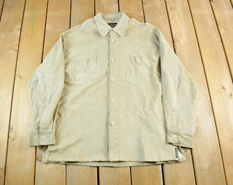 Può includere: Una camicia color beige chiaro con bottoni e due tasche sul petto. La camicia a maniche lunghe ha un colletto e polsini con bottoni. La camicia è realizzata in materiale morbido ed è appoggiata su una superficie di legno.