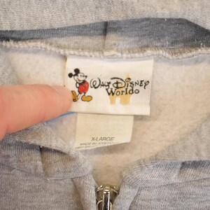 Op de afbeelding: Close-up van een grijze hoodie met ritssluiting en een witte binnenkant. Het label toont het Walt Disney World-logo en een cartoon Mickey Mouse. De maat is X-LARGE.