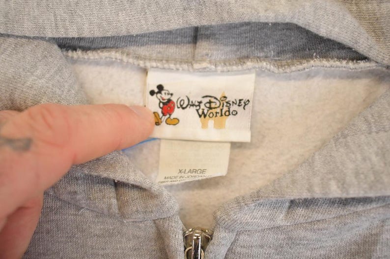 Op de afbeelding: Close-up van een grijze hoodie met ritssluiting en een witte binnenkant. Het label toont het Walt Disney World-logo en een cartoon Mickey Mouse. De maat is X-LARGE.