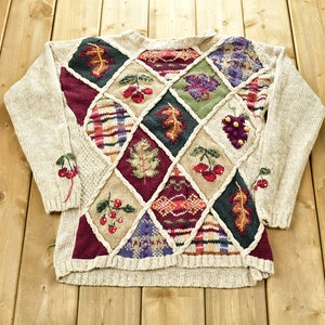 May include: A beige knitted jumper with a diamond pattern featuring embroidered autumnal motifs, including leaves, grapes, and cherries.