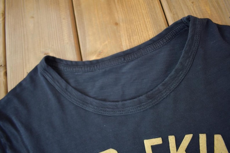 May include: Close-up of the neckline of a dark blue t-shirt with a gold graphic print that reads "EKI".