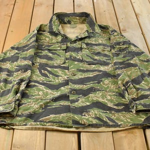 Op de afbeelding: Een overhemd met lange mouwen en knoopsluiting met een tijgerstreep camouflagepatroon in tinten olijfgroen, donkerbruin en zwart. Het overhemd heeft een standaard kraag, twee borstzakken met knopen en manchetten met knopen aan de mouwen.