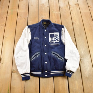 May include: A navy blue and white varsity jacket with leather sleeves. The jacket has a large embroidered patch on the back that says "GENERACION 80-83".