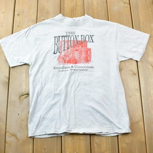 Könnte beinhalten: Weißes T-Shirt mit einer roten und schwarzen Grafik eines Gebäudes mit dem Text "The Button Box Accordions & Concertinas Antiques - New & Used Instruments".