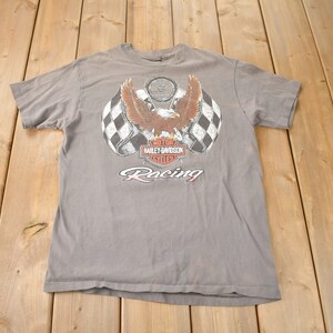 May include: A gray t-shirt with a Harley-Davidson logo and an eagle with outstretched wings. The logo is surrounded by checkered flags and the text "Racing" is below the logo.