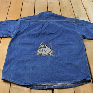 Può includere: Una camicia a maniche corte in denim blu con abbottonatura e un grafico ricamato al centro sul retro. Il ricamo raffigura un personaggio dei cartoni animati stilizzato con capelli scuri, occhiali da sole e labbra pronunciate.