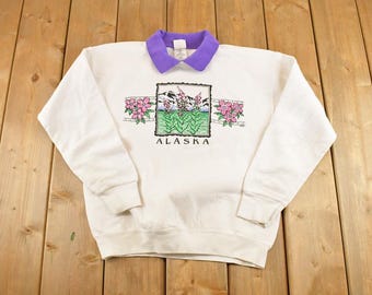 May include: White sweatshirt with a purple collar. The front features a graphic of a mountain scene with pink flowers and the word "ALASKA" in black. The sweatshirt has long sleeves and a ribbed cuff.