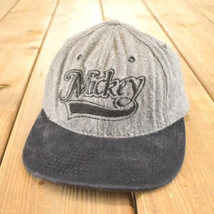 Op de afbeelding: Grijze baseballpet met een zwart vizier. Het woord "Mickey" is in zwart script op de voorkant geborduurd. De pet is gemaakt van een textuurstof en ligt op een houten oppervlak.