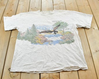 May include: A white short-sleeve t-shirt featuring a graphic print of a bald eagle flying over a waterfall, pine trees, and rocky terrain. The text above the eagle reads "HARRIS STATE PARK" in small, dark green lettering on the front of the shirt.
