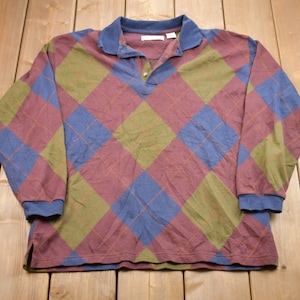 Puede incluir: Polo de manga larga con un estampado de rombos en burdeos, azul y verde oliva. La camisa tiene cuello y puños azules, y una tapeta de tres botones. La camisa está hecha de un material suave y cómodo.